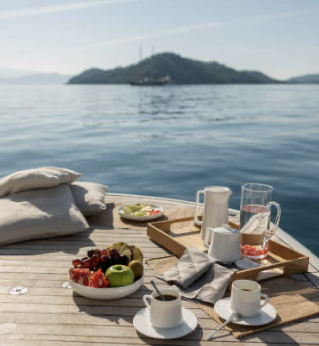 Colazione apparecchiata sul ponte della NOAH 29: caffè, frutta, pasticceria, isolotto sullo sfondo